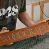 Close-up of a wooden sign engraved with the word “Woodcraft,” showing a smooth resin-filled design. The background shows hands at a workbench, and bold text reads “Beginner Resin Sign.