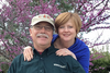 Two people smiling outdoors near a blooming tree, one wearing a Woodcraft shirt and cap.