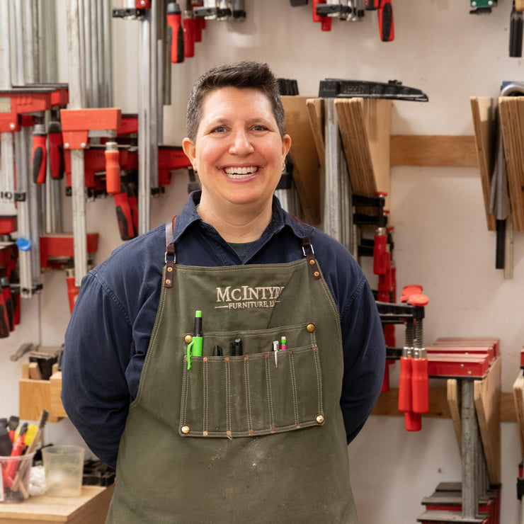 A smiling Kim McIntyre standing in front of a wall of clamps in a woodshop.