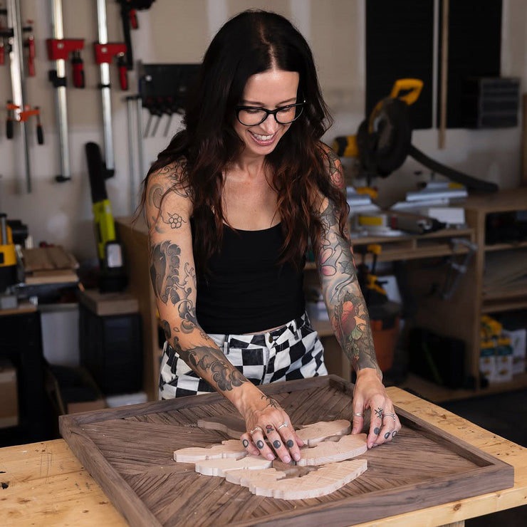 A smiling woman in a wood shop making a do it yourself mosaic on a workbench.