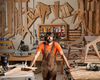 Woodworker in workshop surrounded by wooden templates, tools, and furniture frames.