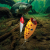 Two hand-turned wooden fishing lures with painted finishes and treble hooks underwater near a largemouth bass.