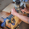 Hands using a router with Leigh B975 box joint jig to cut dovetail joints on wood pieces clamped to a workbench
