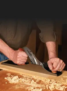 Person using a hand plane on a wooden surface with wood shavings.