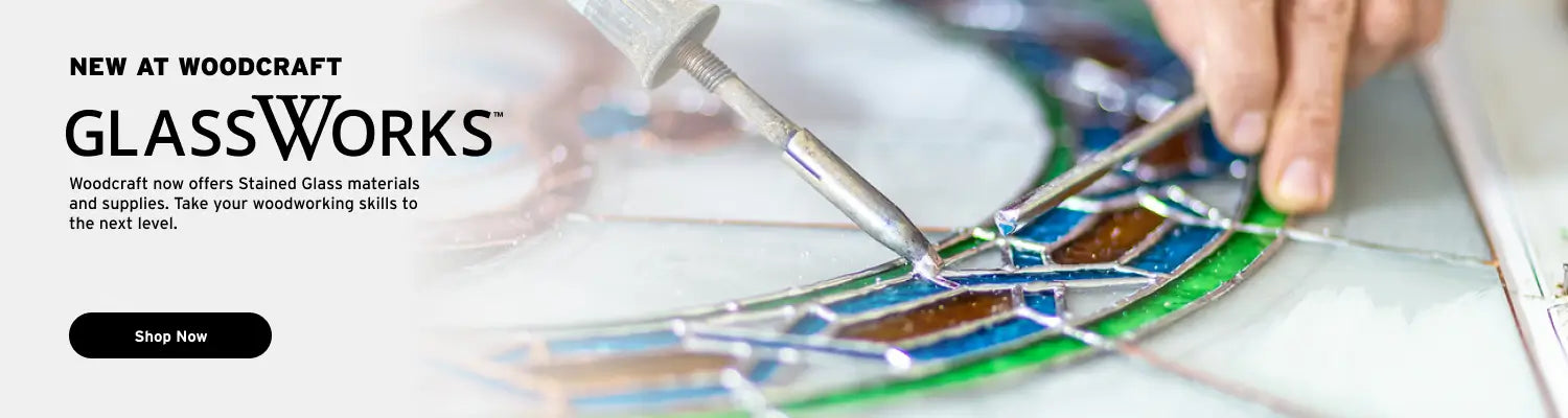 Promotional banner for Woodcraft GlassWorks showing a close-up of a stained glass artist's hand working on a colorful stained glass panel, with text on the left reading 'NEW AT WOODCRAFT GLASSWORKS' and a black 'Shop Now' button.