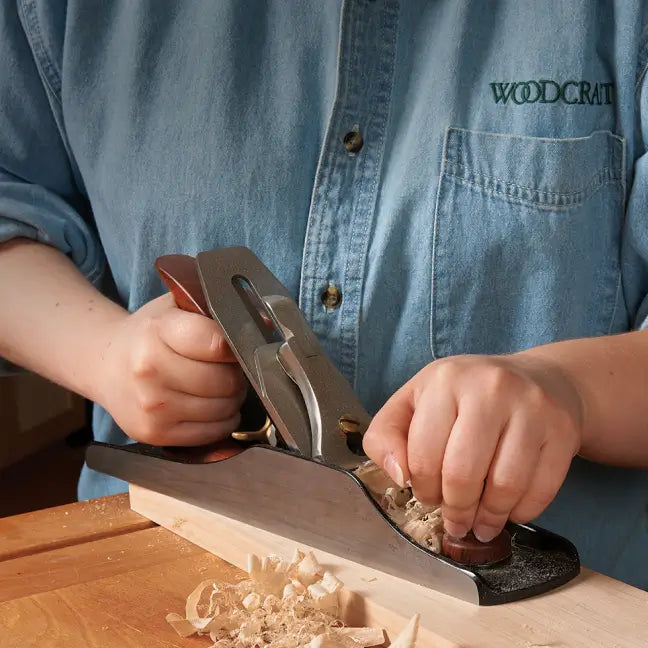 Person using a hand plane on a piece of wood with a blue shirt labeled 'WOODCRAFT'.